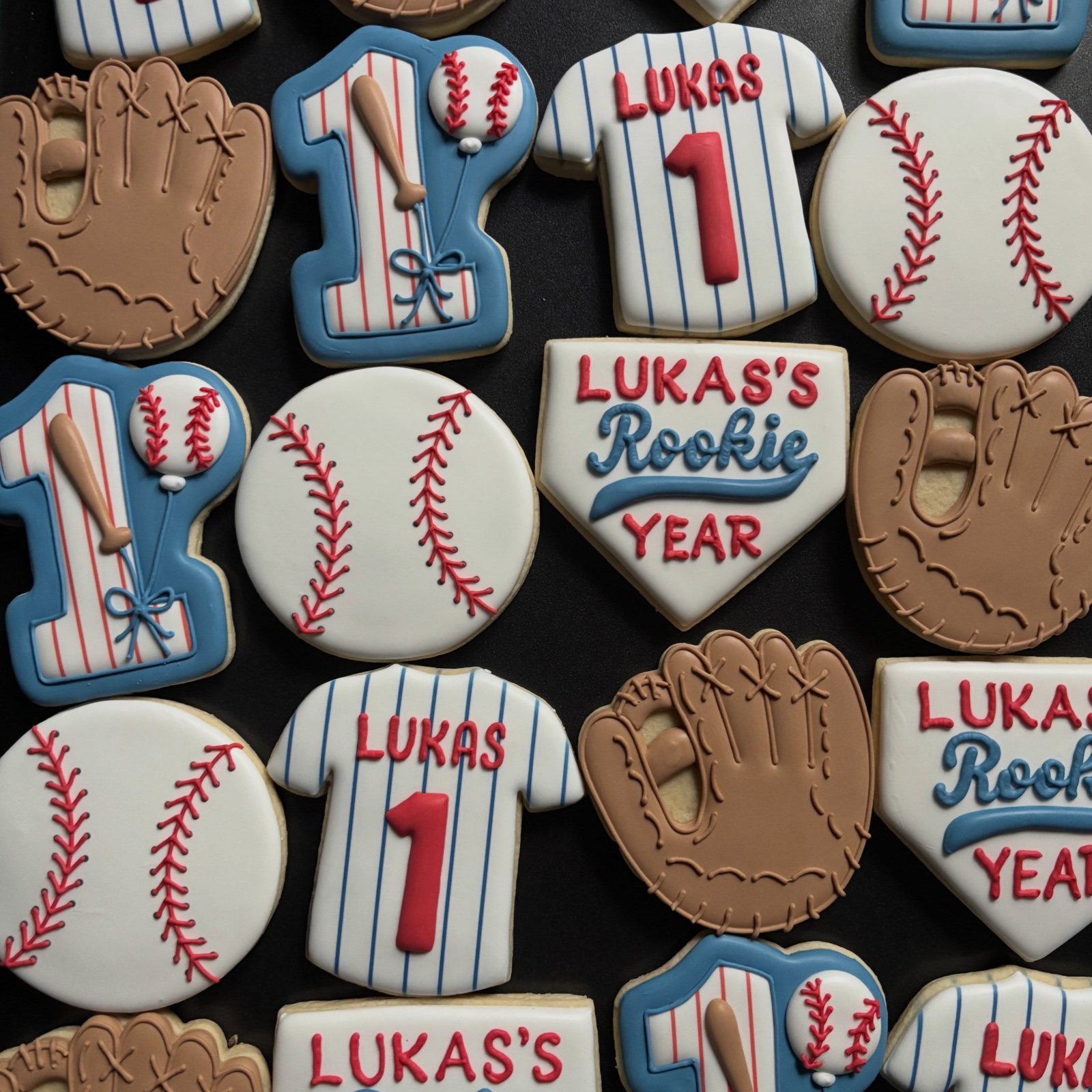 Baseball first-birthday cookies on a black tray: pinstripe jerseys reading Lukas, baseballs, mitts, a number one and a home plate reading Lukas's Rookie Year.